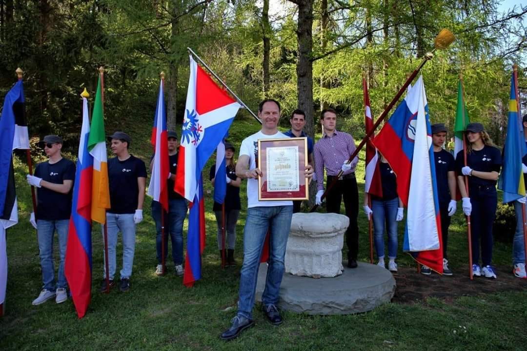 Jože Potrebuješ z zahvalo v rokah ter zastavama Društva Heraldica Slovenica in države Slovenije, skupaj z zastavami vseh držav Evropske unije. GEOSS, 9. 4. 2017.